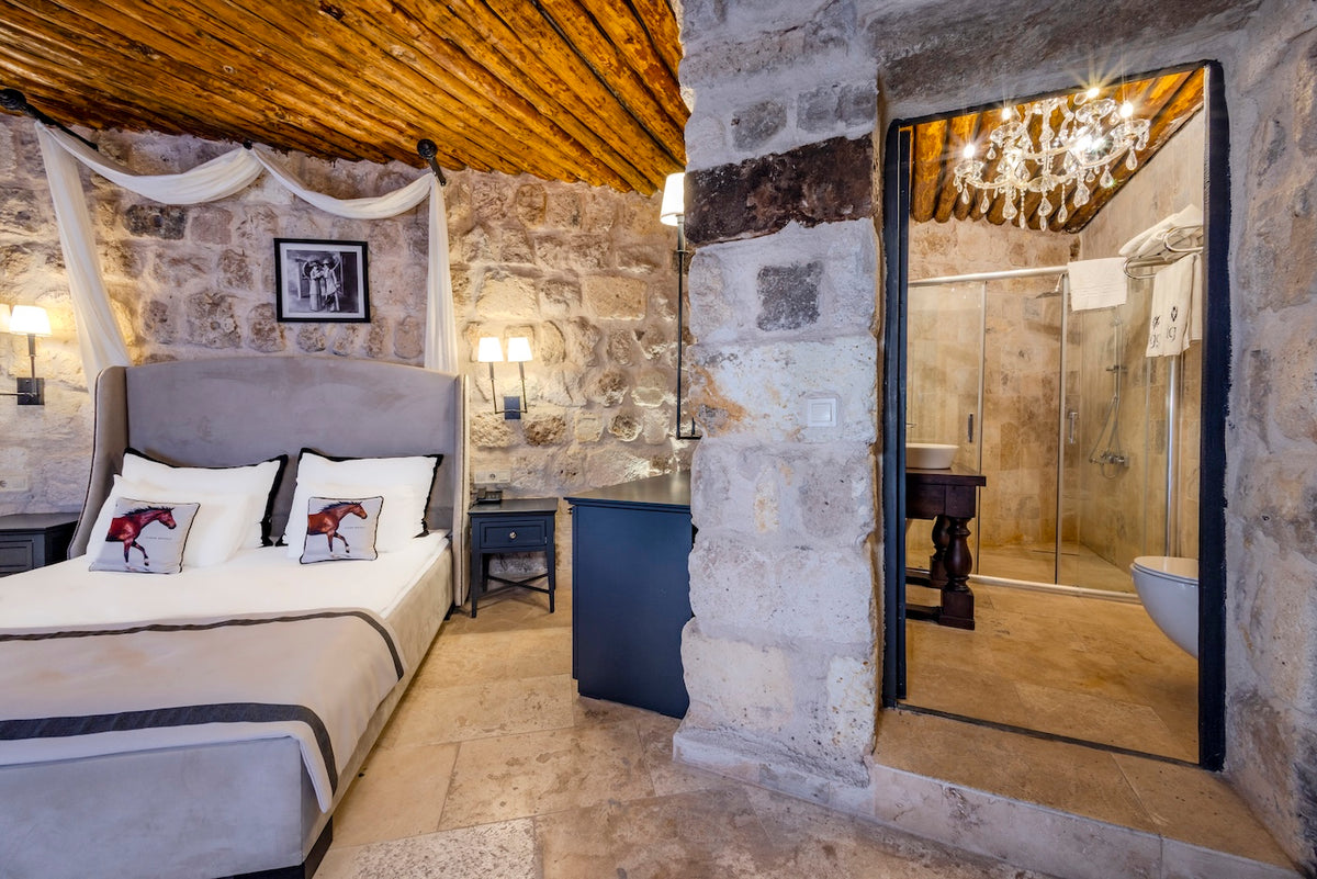 Bedroom with stone walls, bed with decorative pillows, and a bathroom reflection in a mirror.